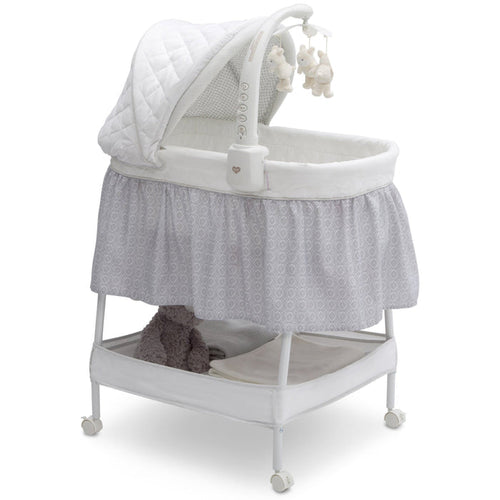 A white Deluxe Gliding Bassinet with a gray patterned skirt, a white mattress pad, and an electronic mobile with plush bears. The bassinet has wheels and a canopy.