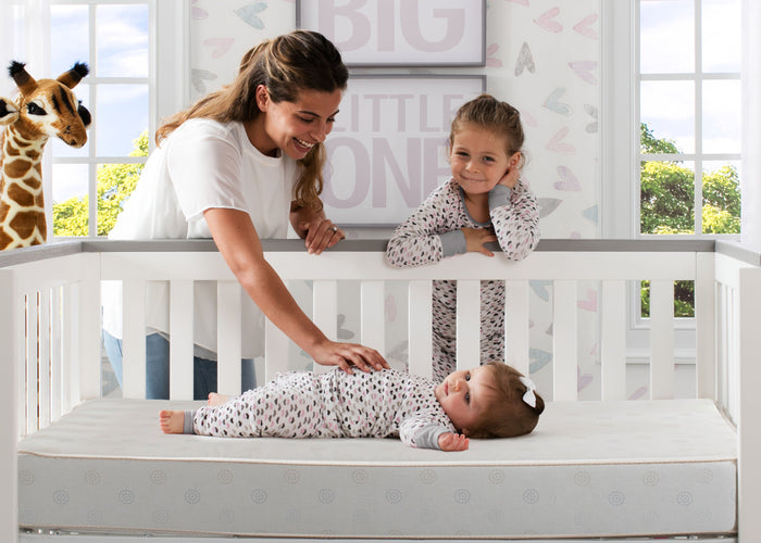 A adult and a child are looking at a baby lying on a dual sided crib and toddler mattress.